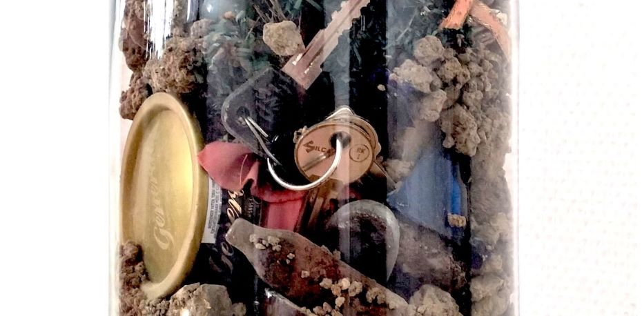 A close-up view of a glass container filled with various objects, including a keyring with keys, a bottle cap, rocks, and other miscellaneous items. The contents appear to be a collection of small, everyday objects, possibly found items or memorabilia.