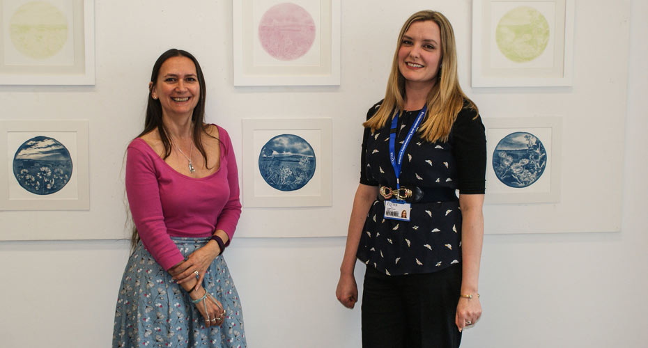 Two women standing in front of a wall displaying framed circular artworks in pastel colors. The woman on the left is wearing a pink top and a patterned skirt, while the woman on the right is wearing a black top with a blue lanyard and dark pants. Both are smiling, creating a welcoming and friendly atmosphere in the gallery setting.
