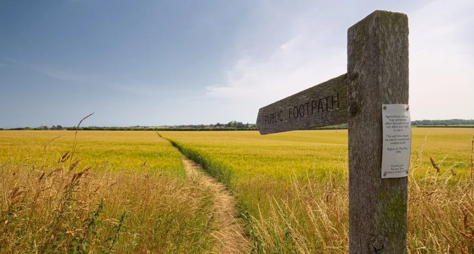 A wooden sign directing towards a path through a grassy field under a clear blue sky.