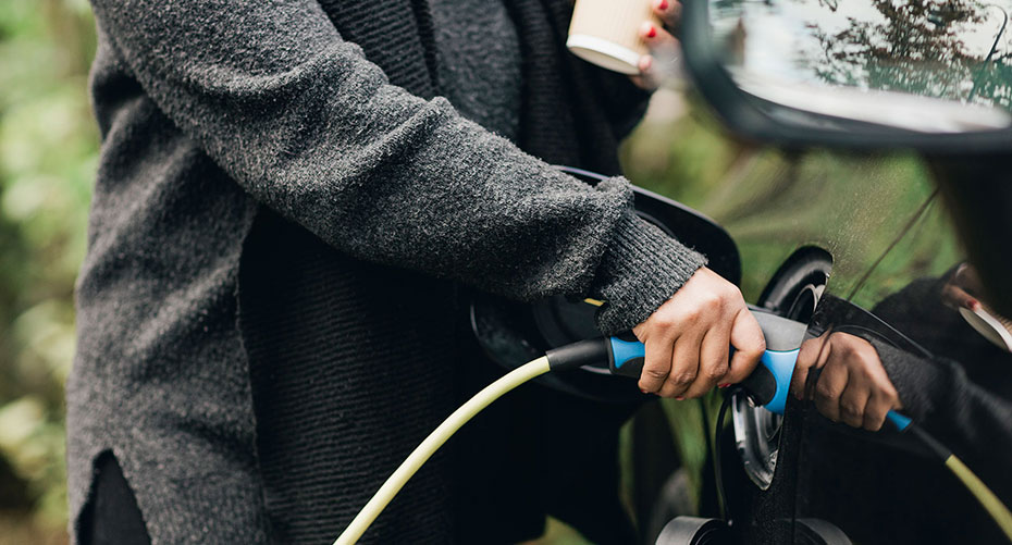 Midsection of mid adult woman charging electric car. She's holding a coffee cup and seems to be in the woods.