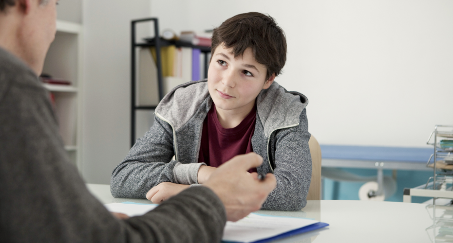 A young person sitting at a table with a person in the background