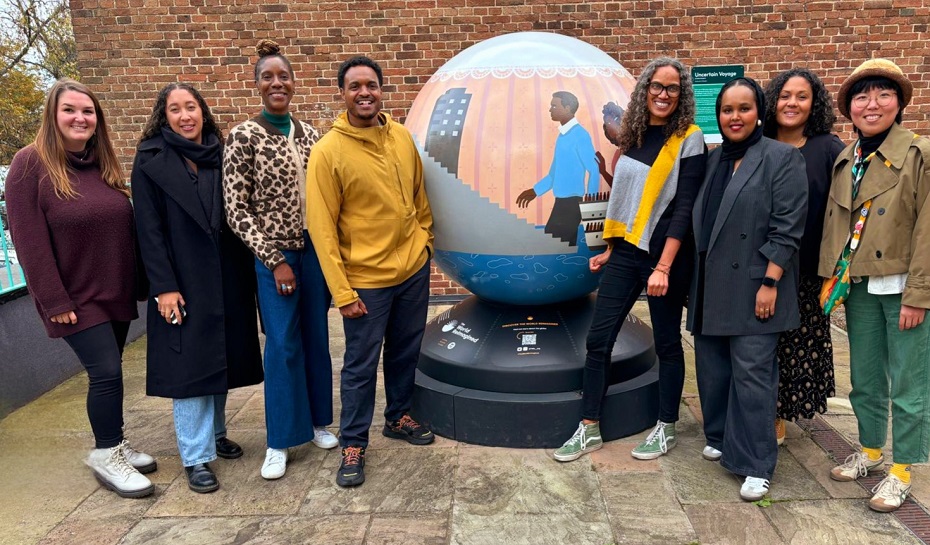 Eight people standing either side of a globe-shaped art installation outside. It features a man and woman who have arrived in Britain from the Caribbean; also visible is the HMT Empire Windrush.