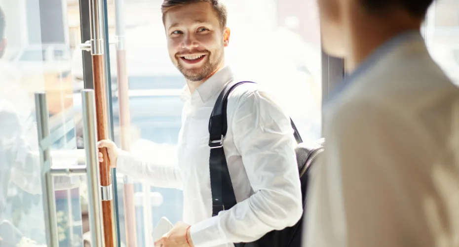 A smiling man stands in front of a glass door at the University of Exeter, showcasing a welcoming atmosphere.